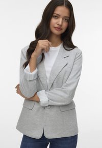 Jasnoszary żakiet z klapami, dwiema przednimi kieszeniami i paskowanymi mankietami. Nosi się go na białym t-shircie i niebieskich dżinsach. Miękka, teksturowana tkanina.