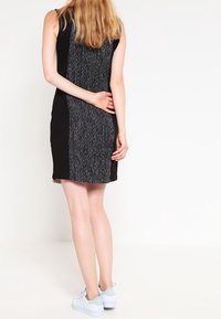 Schwarzes ärmelloses Kleid mit einem strukturierten grauen Panel auf der Rückseite. Das Model trägt weiße Sneakers. Das Kleid hat einen geraden Schnitt und ein minimalistisches Design.