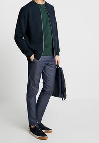 Námořnická texturovaná bomber bunda, zelené tričko s kulatým výstřihem, šedé kalhoty, tmavé tenisky s hnědou podrážkou a taška na rameno, postoj stojící.