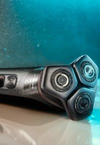 Elektrische scheermes met een zwarte kunststof behuizing, drie ronde scheerkoppen en zilveren accenten. Oppervlak is nat met waterdruppels.