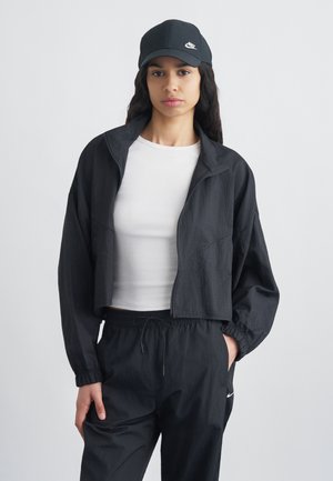 Femeie purtând șapcă neagră Nike, jachetă neagră scurtă, top alb mulat și pantaloni negri cu șnur, stând pe fundal simplu.