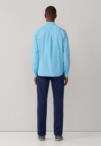 Camisa de botones azul claro con mangas largas, pliegue en la parte trasera y dobladillo redondeado, combinada con pantalones azul marino y calzado azul.