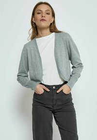 Cardigan grigio chiaro con maniche lunghe, design aperto sul davanti e polsini a costine, abbinato a una camicia bianca e jeans neri a vita alta.