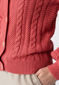 Stickad cardigan i korall med texturerade mönster, med kabeldetaljer och ribbad nederkant. Knappar framtill med en avslappnad passform.