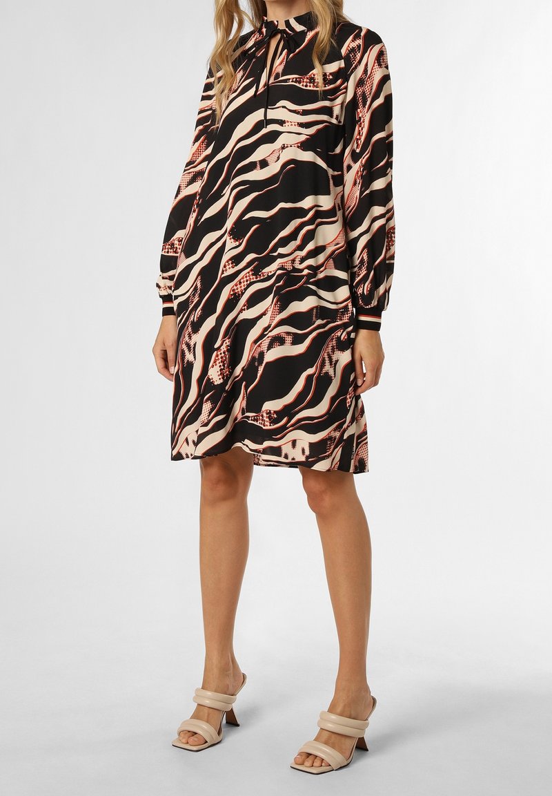 Femme portant une robe longue jusqu'aux genoux à manches longues avec des motifs abstraits ondulés noirs, beiges et rouges, associée à des sandales beiges à talons.