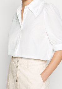 Hvid kortærmet skjorte med en flæsekrave, knaplukning og blødt bomuldsstof, parret med en cremegulv nederdel med forreste knapper.