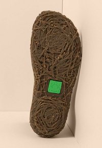 Suela de zapato con un patrón texturizado en forma de ramita y un logo cuadrado verde con una rana en el centro, sobre un fondo beige.