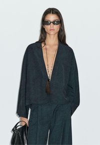 Černá, oversized lněná blůza s širokými rukávy a hlubokým V-výstřihem, doplněná volnými kalhotami ve stejném provedení. Doplňky zahrnují dlouhý náhrdelník a sluneční brýle.