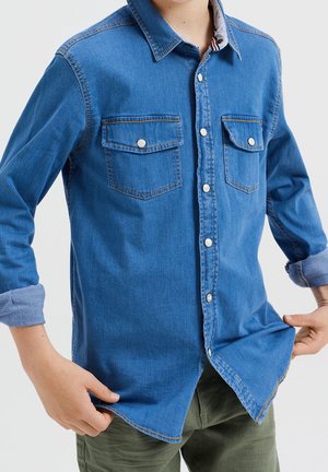 Chemise en denim bleu à boutons, avec deux poches poitrine, quincaillerie argentée et manches retroussées. Texture douce avec une coupe décontractée et un col pointu.