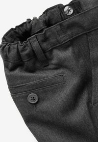 Détail d'un pantalon gris foncé avec taille élastique, poche arrière boutonnée et passant de ceinture sur fond blanc.