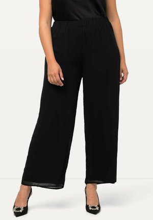 Vrouw die zwarte wijde broek draagt en zwarte puntige hakken met decoratieve gespen, staand tegen een effen achtergrond.