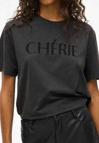 Sort bomulds T-shirt med broderet "CHÉRIE" logo i mørk tråd. Korte ærmer og en løs pasform, kombineret med sorte faux læderbukser.