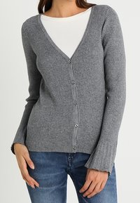 Kvinna i en grå ribbad knäppt cardigan över en vit skjorta, tillsammans med blå jeans, står med händerna vid höfterna.