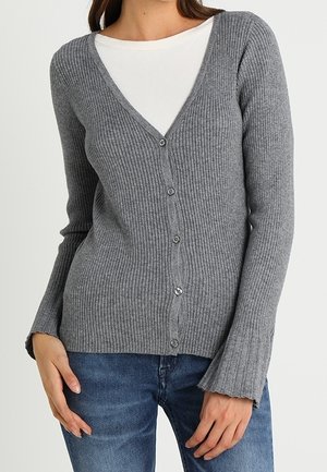 Vrouw in een grijze geribbelde cardigan met knopen over een witte shirt, gecombineerd met blauwe jeans, staand met haar handen bij haar heupen.