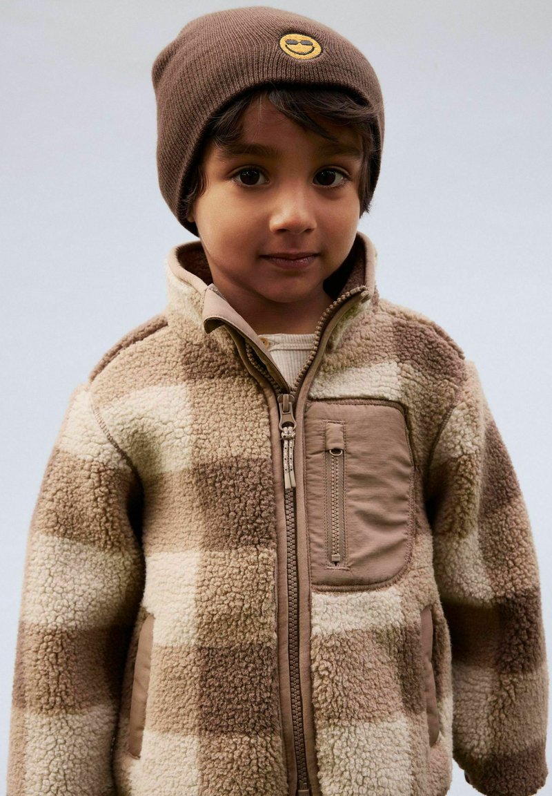 Bambino piccolo che indossa un berretto marrone con toppa a faccina sorridente e una giacca in pile beige a quadri con tasca sul petto con cerniera, rivolto verso l'obiettivo.