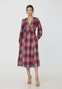 Robe à carreaux rouge et bleu avec des manches longues, décolleté en V profond et poches. Longue jusqu'au sol, avec une taille froncée et un tissu texturé.