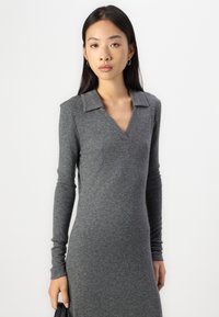 Gray long-sleeve dress with a V-neck and collar. Made of soft textured fabric, form-fitting silhouette. Model holds a small black bag.
