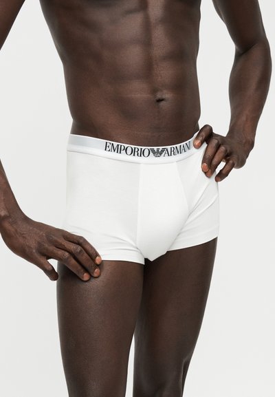 Άνδρας μοντέλο φορώντας λευκά εσώρουχα Emporio Armani, στέκεται με το ένα χέρι στη μέση και το άλλο ακουμπισμένο στον μηρό.