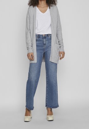 Cardigan grigio chiaro su una t-shirt bianca a scollo a V, abbinato a jeans blu a gamba larga e scarpe con plateau color crema. Include tasche laterali sul cardigan.