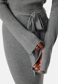 Šedé žebrované pletené šaty s dlouhými rukávy a opaskem. Mají texturovanou tkaninu, přiléhavý střih a detail otevřených rukávů s mašlí v pase.