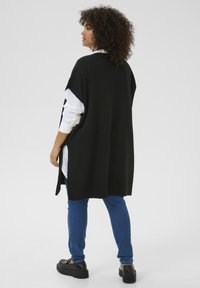 Czarny, oversize sweter z bocznymi rozcięciami, noszony na białej koszuli, w połączeniu z niebieskimi dżinsami i czarnymi butami na platformie, z teksturowanej tkaniny.