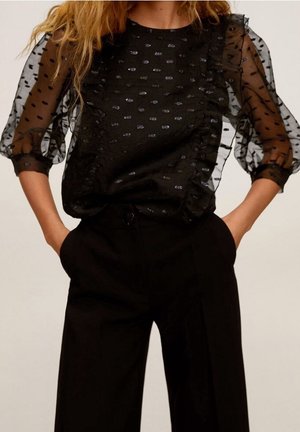 Femme portant un chemisier noir transparent à pois avec des volants et un pantalon large noir taille haute, les mains dans les poches.