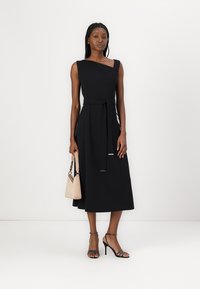 Robe noire sans manches avec un décolleté en diagonale, une ceinture à la taille et une jupe fluide. Le modèle tient un sac à main beige clair et porte des talons à lanières.