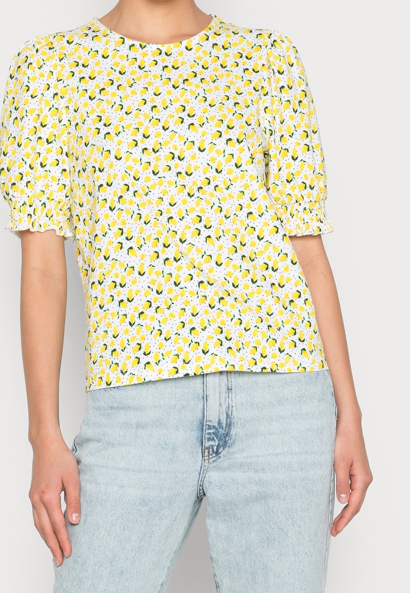 Kobieta ubrana w białą bluzkę w żółty nadruk cytryn oraz bufiaste krótkie rękawy, zestawioną z jasnoniebieskimi, wysokimi dżinsami.