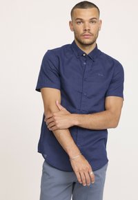 Chemise à manches courtes bleu marine à boutons avec une finition texturée, dotée d'un col pointu et d'un logo discret sur la poche poitrine.