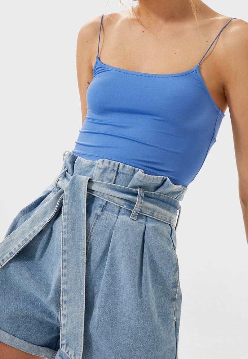 Femme portant un haut bleu ajusté à bretelles spaghetti et un short paperbag en denim clair taille haute avec une ceinture nouée.
