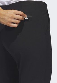 Calças pretas ajustadas com uma textura suave, apresentando um bolso traseiro e uma mão a colocar um objeto no bolso.