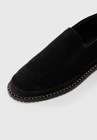 Chaussure noire en suede à enfiler avec des coutures beiges visibles autour de la semelle et un panneau élastique sur les côtés, sur un fond blanc.