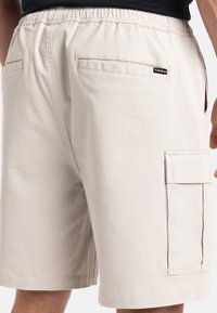Pantalones cortos cargo beige con cintura elástica, dos bolsillos traseros y un bolsillo lateral cargo con solapa. El material parece suave y duradero.