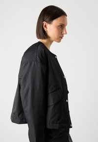 Černá lehká bunda s mírně zkráceným střihem, uzavíráním na patentky a dvěma předními kapsami. Hladká textura s minimalistickým stylem.
