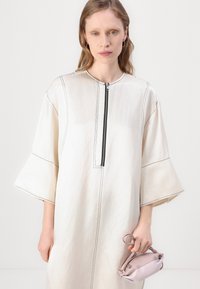 Krémové šaty s čiernym šitím, polovičné rukávy a zips na uzatvorenie. Modelka drží malú ružovú kabelku. Hladká textúra.