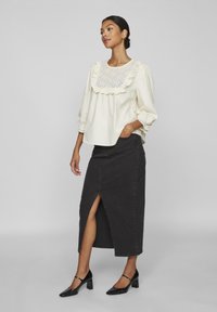 Blouse en coton blanc avec un col en volants bordé de dentelle, des manches ballon, associée à une jupe en denim noir avec une fente à l'avant. Talons noirs.