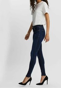 Jean skinny en denim bleu foncé avec une taille haute et un design à cinq poches, porté avec un t-shirt blanc uni et des talons hauts noirs.