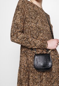 Kvinna i långärmad klänning med bruna och svarta prickar som håller en liten svart väska i krokodilmönster med guldfärgad hårdvara.