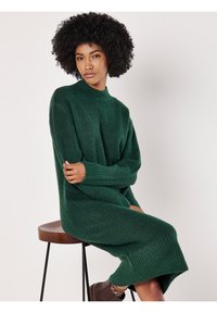 Grünes Strickkleid mit hohem Kragen, langen Ärmeln und gerippten Bündchen und Saum, aus einem strukturierten Stoff, getragen über braunen Knöchelstiefeln.