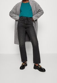 Cappotto grigio lungo su una maglietta teal, abbinato a jeans neri a vita alta e mocassini neri con dettagli in hardware dorato.