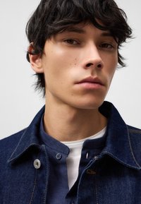 Giacca di denim di colore blu scuro, con bottoni in metallo argentato e cuciture arancioni, indossata sopra una maglia henley blu navy con scollo rotondo.