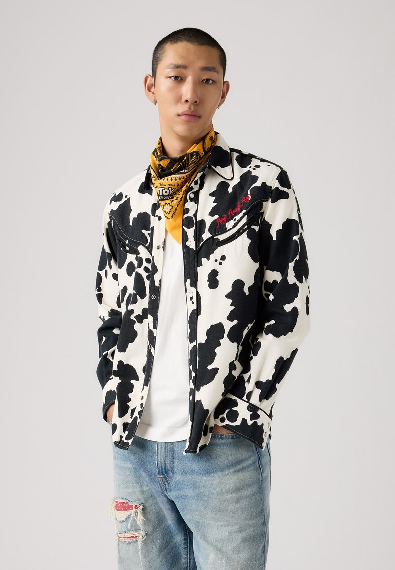 Kuhmønstret denimskjorte i sort og hvit, med frontlommer, kontrasterende sømmer, parret med slitt blå jeans og et gult bandana.