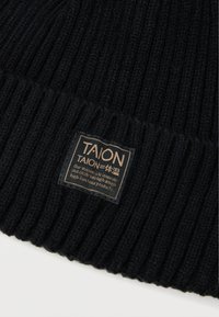 Beanie nero a coste con tessuto texturizzato. Presenta una patch rettangolare in tessuto con il logo "TAION" e la dichiarazione di missione in beige.