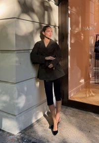 Giacca nera doppiopetto con maniche larghe, abbinata a pantaloni neri a vita alta e tacchi a punta neri, in piedi contro un muro di colore chiaro.