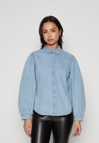 Lyseblå denimskjorte med krage, knappefront og puffermer. Har sømmer med stikninger og en buet kant.