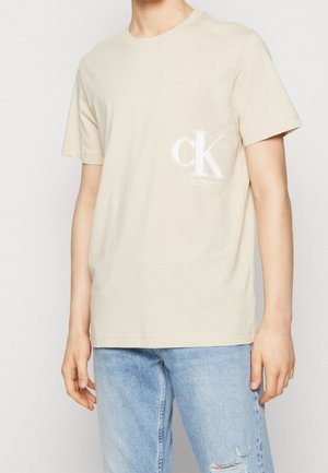 Osoba nosząca beżowy t-shirt z krótkim rękawem z białym logo "cK" na piersi oraz jasnoniebieskie podarte dżinsy.