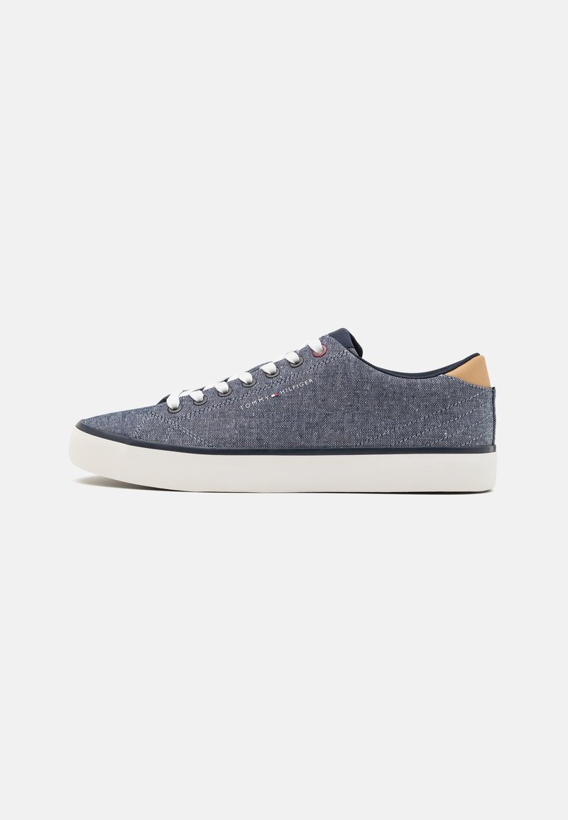 Basket basse en tissu denim avec lacets blancs, languette de talon beige, bordure marine et semelle blanche en caoutchouc sur fond uni.