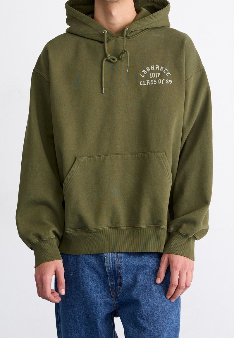 Oliwkowa bluza z kapturem wykonana z mieszanki bawełny, z przednią kieszenią i sznurkiem w kapturze. Na klatce piersiowej napis "Carhartt Klasa 89".