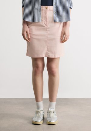 Nainen, joka pukeutuu vaaleanpunaiseen nappihameeseen, raidalliseen paitaan, valkoisiin sukkiin ja hopeisiin lenkkareihin, seisoo harmaalla lattialla valkoista taustaa vasten.