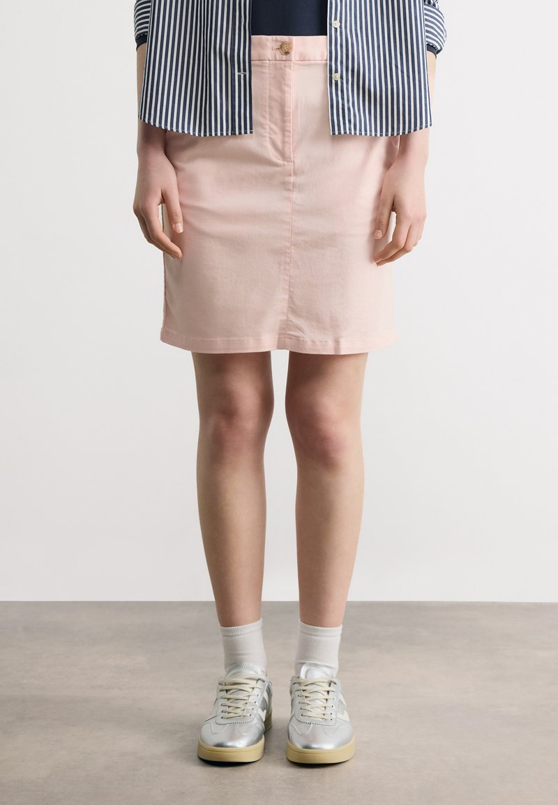 Ženska, oblečena v roza krilo z gumbi, črtasto srajco, bele nogavice in srebrne superge, stoji na sivem tlaku pred belim ozadjem.